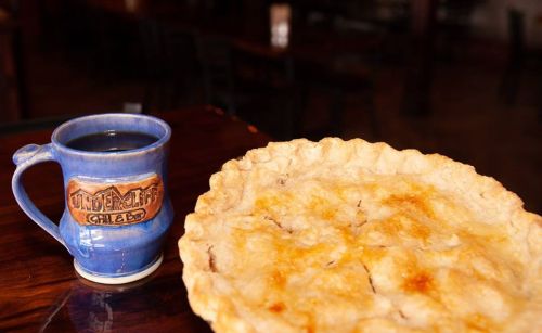 A slice of golden-brown pie next to a blue mug of coffee on a wooden table.