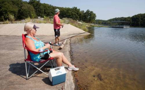 A woman sits in a chair by the water, fishing, while a man stands nearby, also fishing, on a sunny day.