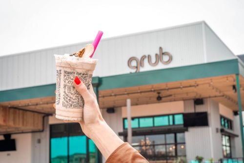 A hand holds a milkshake with a pink straw in front of a restaurant sign that reads "grub."