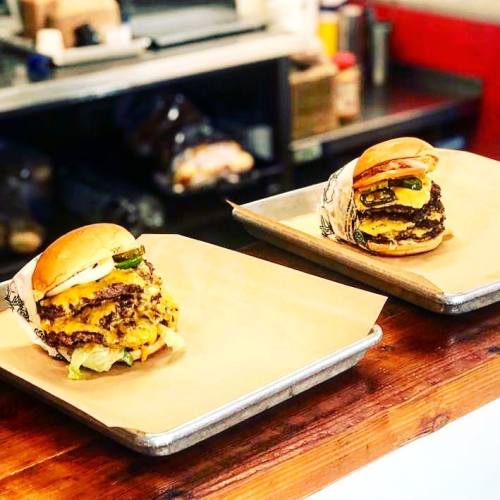 Two stacked cheeseburgers on trays, featuring layers of beef, cheese, lettuce, and pickles, set against a kitchen backdrop.