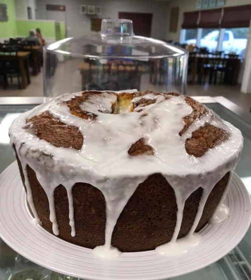 A round cake with a glossy white icing drizzled on top, displayed under a glass dome in a café setting.