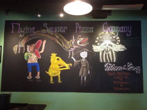 Chalkboard mural featuring whimsical characters and the text "Flying Saucer Pizza Company" with pizza slice options.