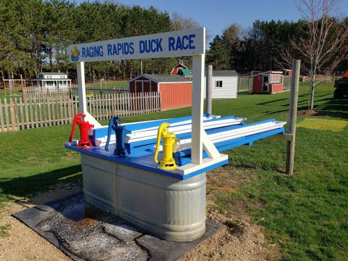 A colorful duck race setup with water channels and pumps, surrounded by green grass and small buildings in the background.