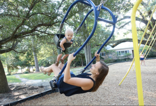 A woman swings while a toddler playfully climbs on the swing set in a sunny park. Trees and a house are in the background.