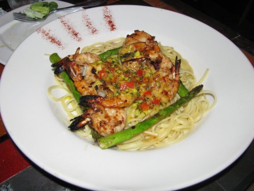 A plate of pasta topped with grilled shrimp, asparagus, and diced tomatoes, garnished with seasoning.