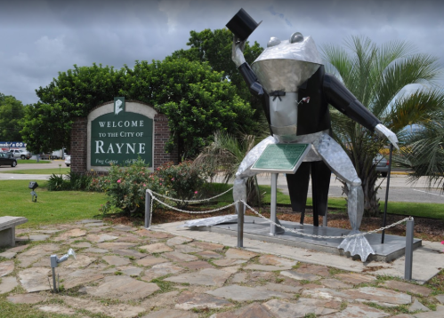 A large frog statue in a tuxedo and top hat stands near a welcome sign for the city of Rayne, surrounded by greenery.