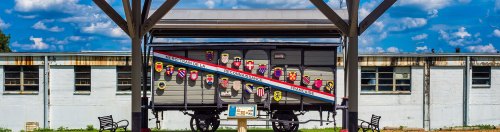 A historic train car displayed under a shelter, featuring colorful shields and a backdrop of blue skies and clouds.