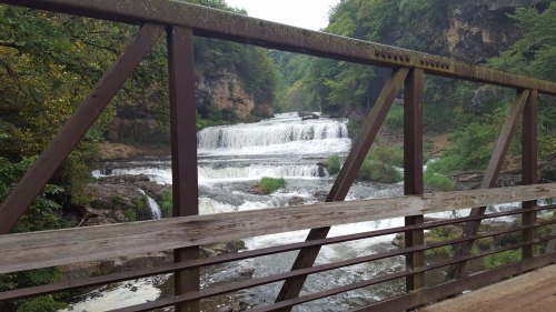 A scenic view of a waterfall framed by a rustic metal bridge, surrounded by lush greenery.