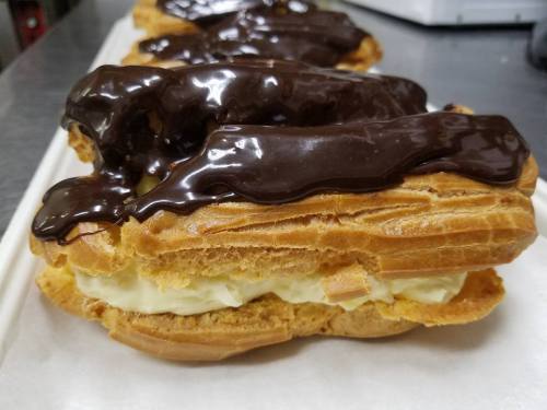 Close-up of a chocolate-covered eclair filled with creamy custard, resting on a white surface.