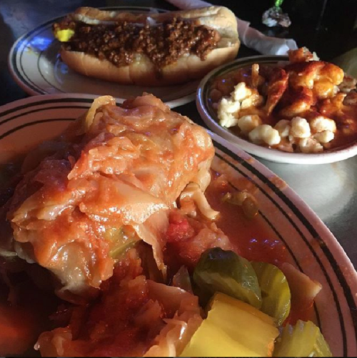 A plate featuring stuffed cabbage rolls, pickles, and a side of macaroni and cheese, with hot dogs in the background.