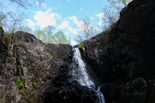 A waterfall cascading down rocky cliffs, surrounded by greenery and a bright blue sky with scattered clouds.