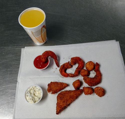 A plate of fried seafood, including shrimp and fish, with dipping sauces and a cup of orange drink.