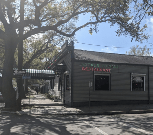 A green and gray restaurant building with "Pascal's Manale" sign, surrounded by trees on a sunny day.