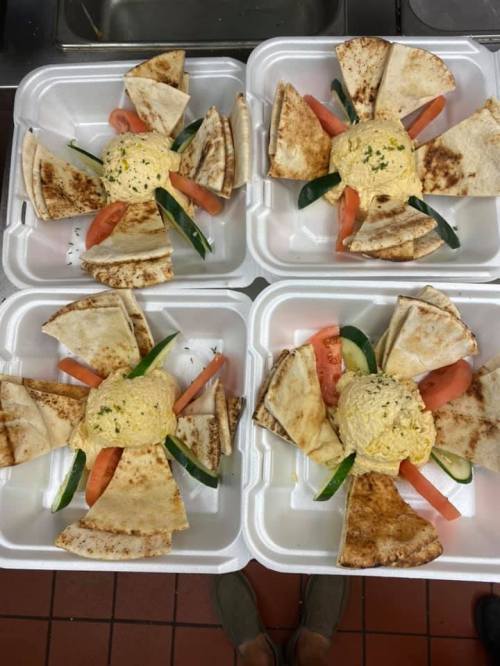 Four takeout containers with pita bread, hummus, and fresh vegetables arranged in a star pattern.