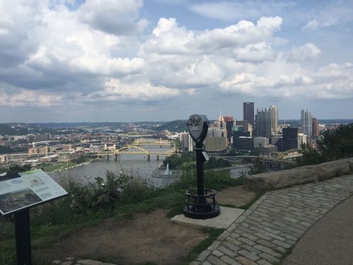 View of Pittsburgh skyline from a lookout point, featuring a viewing telescope and a river with bridges under a cloudy sky.
