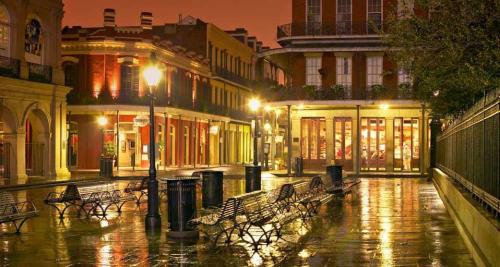 A quiet, illuminated street in New Orleans at dusk, with wet pavement and empty benches under warm streetlights.