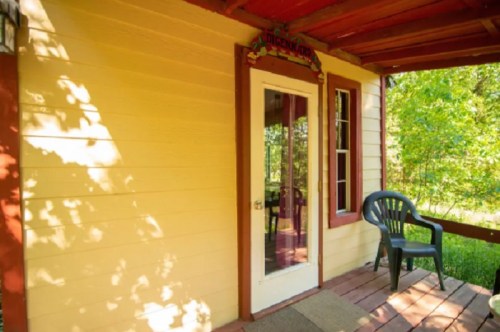 A cozy porch with a chair, featuring a door and windows, surrounded by greenery. Sunlight casts shadows on the wall.