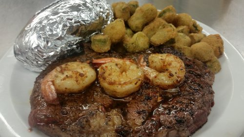 A plate featuring a grilled steak topped with shrimp, a baked potato wrapped in foil, and fried okra.
