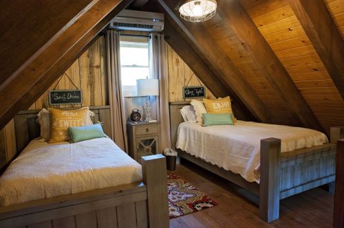 Cozy attic bedroom with two beds, wooden walls, a lamp, and decorative pillows, creating a warm, inviting atmosphere.