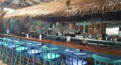 A colorful bar with a thatched roof, featuring blue and teal stools and a well-stocked shelf of liquor bottles.