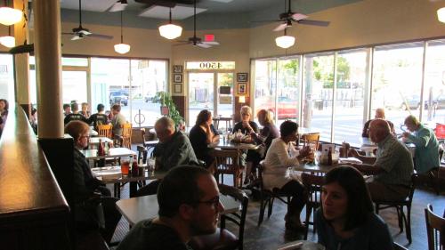 A bustling restaurant interior with diners at tables, large windows, and a warm, inviting atmosphere.