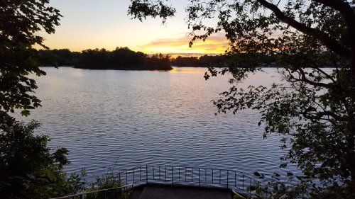 A serene lake at sunset, with calm waters reflecting the colorful sky and surrounded by trees.