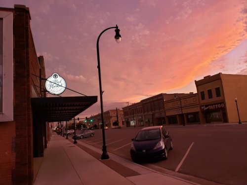 A quiet street at sunset, featuring a shop sign, parked car, and colorful sky with clouds.