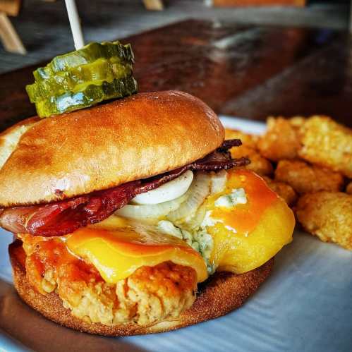A close-up of a chicken sandwich topped with cheese, pickles, and onions, served with a side of tater tots.