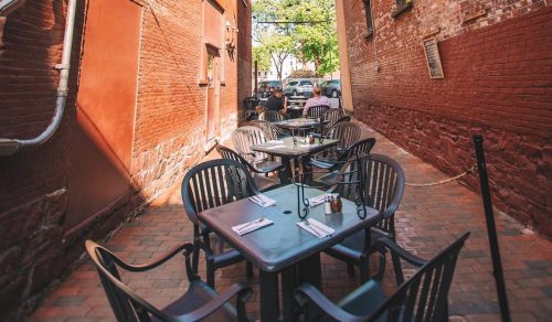 Narrow alley with brick walls, outdoor tables, and string lights under a blue sky.