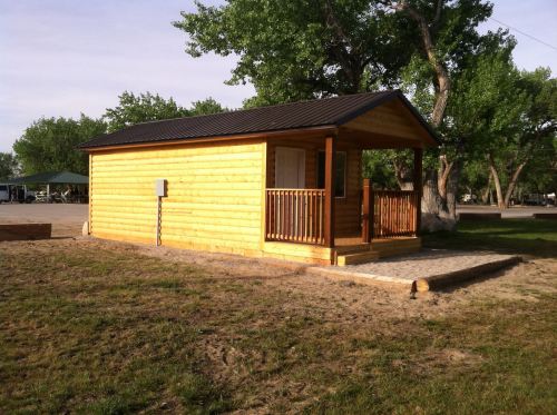 A small wooden cabin with a porch, surrounded by grass and trees, under a clear sky.