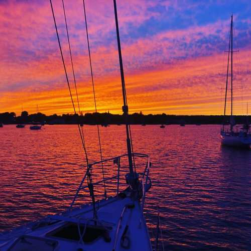 A vibrant sunset over a calm harbor, with sailboats silhouetted against colorful skies and water reflections.