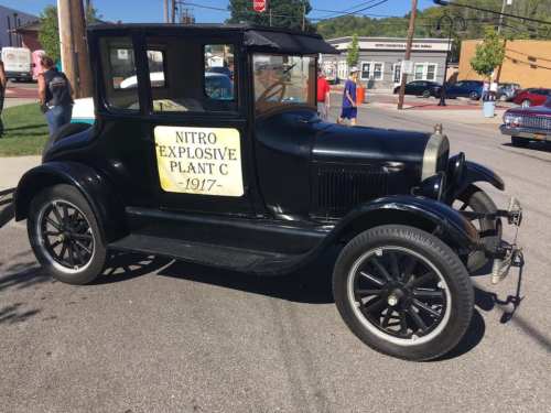 A vintage black car with a sign reading "NITRO EXPLOSIVE PLANT C 1917" parked on a street during an event.