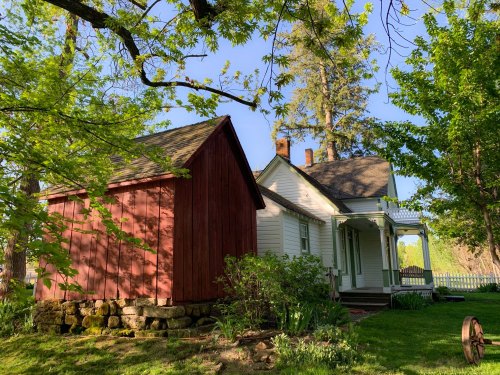 A quaint white house with a red barn, surrounded by lush greenery and trees under a clear blue sky.