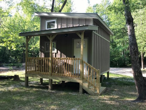 A small wooden cabin with a porch, surrounded by trees in a natural setting.