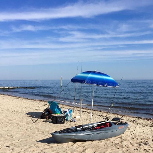 A kayak and fishing gear under a blue umbrella on a sandy beach by the calm sea, with a clear blue sky above.