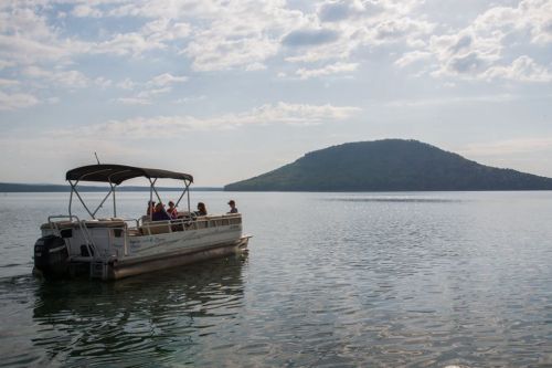 A pontoon boat with passengers floats on calm water, with a distant hill under a cloudy sky.