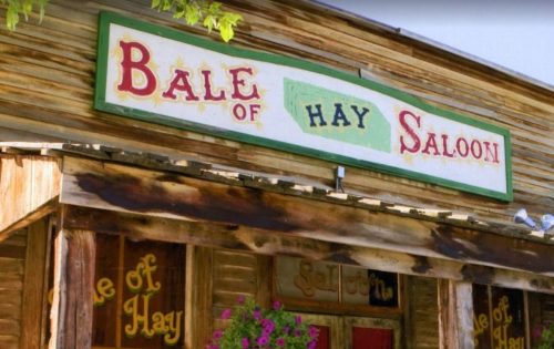 Sign for "Bale of Hay Saloon" on a rustic wooden building with colorful lettering and hanging flowers.