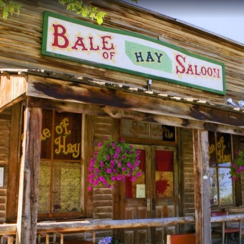 A rustic wooden saloon with a colorful sign reading "Bale of Hay Saloon" and flower baskets hanging by the entrance.
