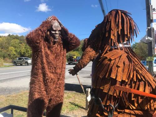 Two Bigfoot figures, one in a furry costume and the other made of rusted metal, stand near a roadside in a sunny setting.