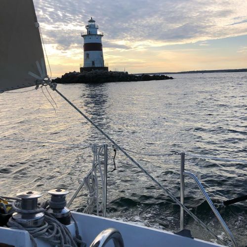 A sailboat's view of a lighthouse at sunset, with calm waters and a cloudy sky in the background.