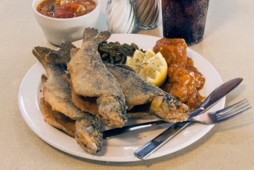 A plate of fried fish with lemon, green beans, sweet and sour sauce, and a bowl of soup, alongside a glass of cola.