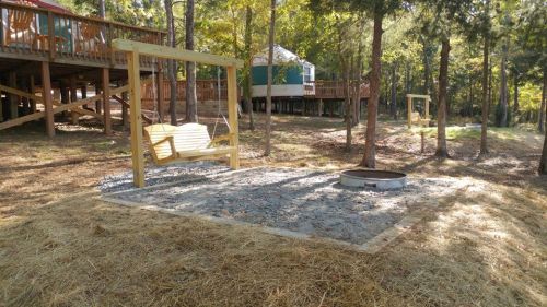 A wooden swing sits on a gravel area surrounded by trees, with a fire pit nearby and a yurt in the background.