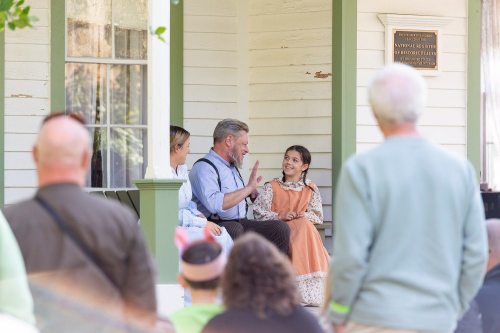 A man in period clothing talks animatedly with two girls on a porch, while a crowd watches from a distance.