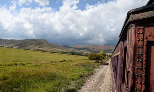 A scenic view from a train, showcasing green fields and mountains under a cloudy sky.