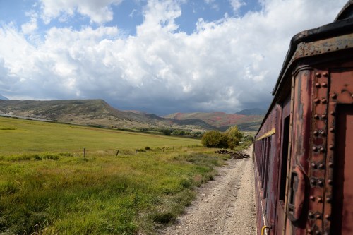 A scenic view from a train, showcasing green fields and mountains under a cloudy sky.