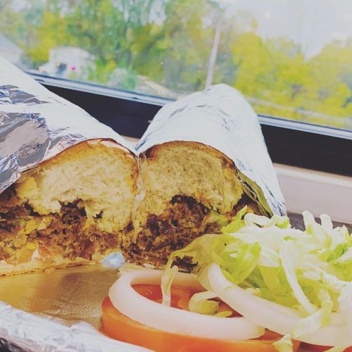 A close-up of a sandwich wrapped in foil, filled with meat and served with lettuce, tomato, and onion on the side.