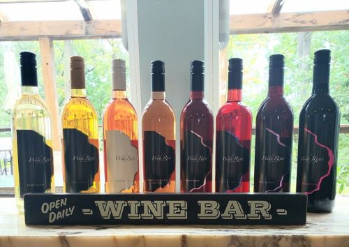 A display of eight wine bottles in various colors, with a sign reading "Open Daily - Wine Bar" in front.