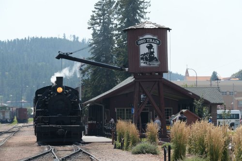 A vintage steam train at a station with a water tower, surrounded by trees and a clear sky.