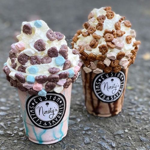 Two colorful milkshakes topped with whipped cream and cereal, one with pastel colors and the other in chocolate.