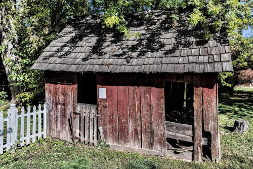 A weathered red shed with a wooden roof, surrounded by greenery and a white picket fence.